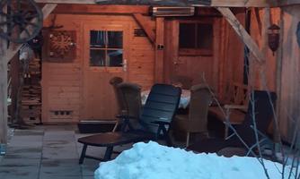 A cozy wooden sauna with a sign above the door. Snow surrounds the sauna, and there are a few chairs in the outdoor area.