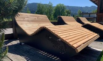Modern designed loungers made of wood are placed on a wooden terrace. In the background, trees and mountains can be seen.