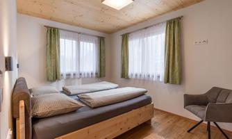 A cozy bedroom with a double bed and wooden floor. The windows have light curtains and the walls are in neutral colors.