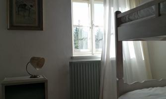 A cozy bedroom with a bunk bed and a window through which daylight streams in. Next to the bed is a nightstand with a lamp.