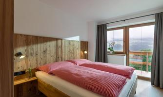 A cozy bedroom with a double bed and a red-and-white plaid pattern. Large windows offer a view of the surrounding nature.