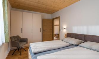 A modern bedroom with a double bed and a chair. The walls are light and the wooden ceiling creates a cozy atmosphere.