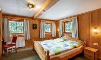 A cozy bedroom with wooden decor and a large bed. The windows offer a view of nature, and there is a chair as well as a bedside table in the room.