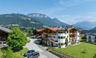 A picturesque landscape with mountains in the background and modern residential buildings. Green meadows and parked cars complete the picture.