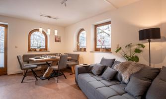 A bright living room with a gray sofa and a dining table. Large windows let in plenty of daylight, and plants provide a homely atmosphere.