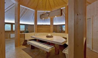 A cozy dining area with a large wooden table and benches. The walls are made of wood, and large windows let in plenty of light.