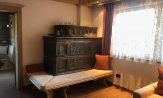 A cozy living space with a historical metal stove and a wooden bench. Brightly illuminated by the daylight streaming through the window.