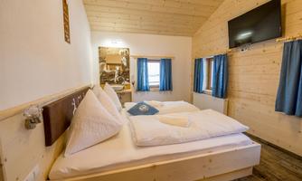 A cozy bedroom with wooden paneling and a double bed. There is a television on the wall and blue curtains at the windows.