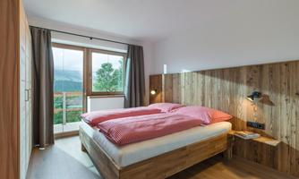 A modern bedroom with wood paneling and a large window. The bed has red pillows and the view shows a green landscape.