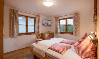 A cozy bedroom with a double bed, striped pillows, and wooden floor. Large windows provide plenty of daylight and a relaxed atmosphere.