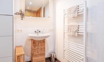 A modern bathroom with a hand basin and a large mirror. Towels hang on a white towel rack and there is a wooden shelf next to it.