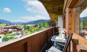 A balcony overlooking green hills and traditional houses. On the table are flowers and breakfast dishes.