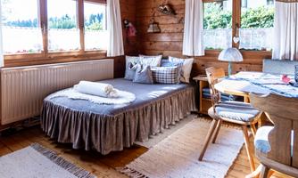 A cozy room with wood paneling and large windows. In the foreground, there is a bed, a table, and some chairs.