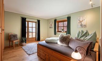 A cozy bedroom with green walls and wooden floor. There is a daybed with cushions and an open door that leads to a view.