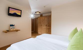 A cozy room with two beds and light green pillows. In the background, a TV and a wooden tower can be seen.