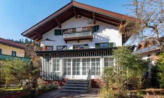 A charming house in alpine style with several windows. The garden is well-maintained and surrounded by trees.