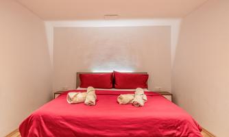 A cozy bedroom with a large red bed. The walls are plain and bright, and several towels are lying on the bed.