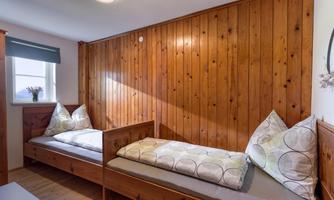 A cozy room with two wooden beds and cushions. The walls are made of wood, and a window lets in daylight.