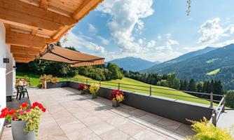 A sunny terrace with flower boxes and a view of the mountains. In the background, green hills and a blue sky can be seen.