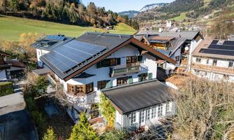 A charming house in a mountainous setting with solar panels on the roof. Surrounded by trees and gentle hills.