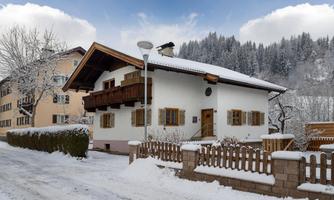 A cozy house in the snow with a traditional roof and wooden windows. The surroundings are filled with trees and snow-covered hills.