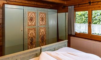A cozy bedroom with wooden paneling and a decorative wardrobe. Large windows let in plenty of daylight.