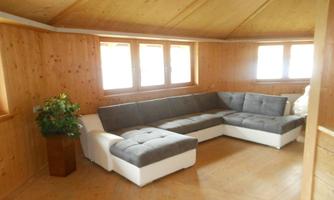 A bright room with a cozy gray corner couch and wooden walls. Large windows let in plenty of light and create a warm atmosphere.