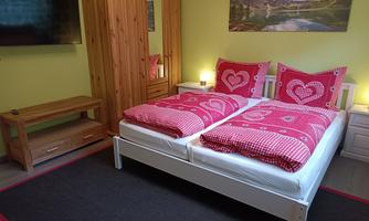 A cozy bedroom with two beds and red-and-white bedding. A picture of a landscape hangs on the wall, and there is a television and a wooden wardrobe.