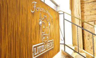 A wooden sign with the inscription "Ferienbauernhof Litzl & Hof". It features a stylized cow motif and is attached to a staircase.
