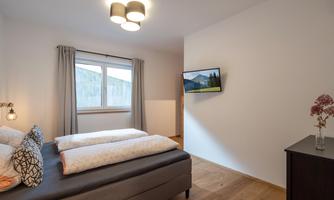 A cozy bedroom with two beds and a TV on the wall. Large windows let in plenty of light, and the room has a modern wooden floor.