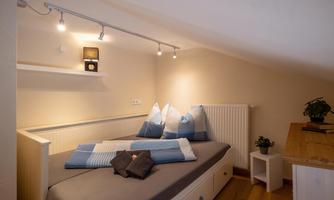 A cozy bedroom with a bed, blue pillows, and soft lighting. Next to the bed is a small table with a plant.