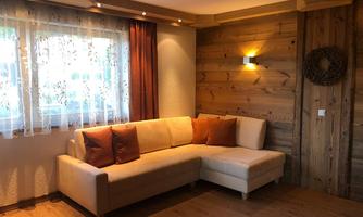 A cozy living room with a beige couch and orange cushions. The walls are made of wood, and a window with curtains lets in plenty of light.