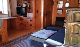A cozy living room with wood paneling and a large window. The furnishings include a sofa, a table, and shelves with books.