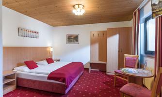 A cozy hotel room with a double bed, red accents, and wooden details. There are chairs and a table as well as a wardrobe in the background.