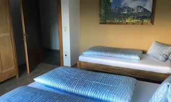A simple guest room with two beds and blue bedding. In the background, there is a landscape painting featuring mountains.