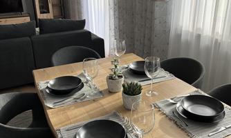 A modern dining area with a wooden table, four chairs, and elegant plates. Stylish lights hang above the table, and a plant arrangement adds a fresh atmosphere.