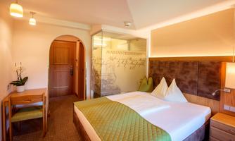 A modern hotel room with a large bed and elegant furnishings. The walls are light, and there is a small workspace as well as plants for a pleasant atmosphere.