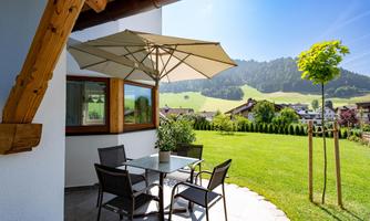 A cozy terrace with a table and chairs, covered by a large sun umbrella. In the background, green meadows and gentle hills can be seen.