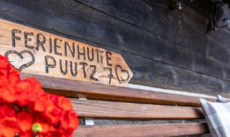 A wooden sign with the inscription "Holiday Cabin Putz" stands against a wall. Red flowers and a cozy bench complement the rustic atmosphere.
