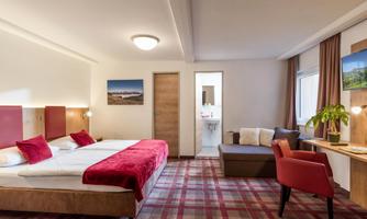 A modern hotel room with two beds, a desk, and a cozy armchair. There is a window with a view of the surroundings and decorative pictures on the walls.