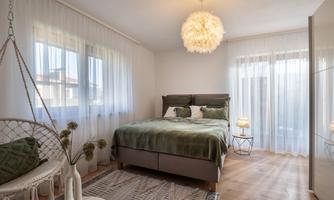 A cozy bedroom with a large bed and a bright, friendly atmosphere. The windows are adorned with white curtains and there is a stylish hanging chair corner.