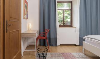 A cozy room with a bed, a desk, and a red chair. Large windows with curtains let in plenty of light.