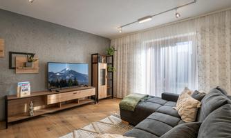 A modern living room with a large sofa, a TV, and a shelf. Large windows provide plenty of daylight.