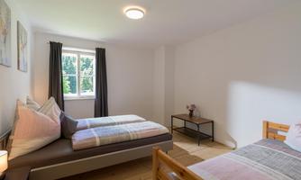A simple room with two beds and a window that lets in a lot of daylight. The walls are light-colored, and there is modern furniture.