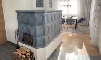 A stylish tiled stove in a bright room. In the background, a dining table and chairs are visible.