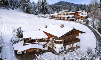 A charming chalet in the snow, surrounded by snow-covered trees. In the background, more houses and gentle hills can be seen.
