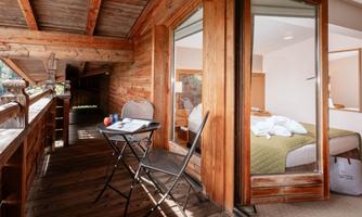 A cozy balcony with a small table and two chairs. In the background, there is a room with a bed and natural wood interior.