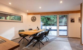 A modern dining room with a large wooden table and black chairs. Large windows offer a view of the green surroundings.