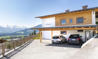 A modern house with a large parking lot and two cars. In the background, the mountains and a clear blue sky can be seen.