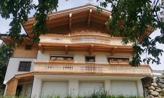 A typical Alpine wooden house with multiple balconies and flower boxes. Surrounded by green grass and trees.